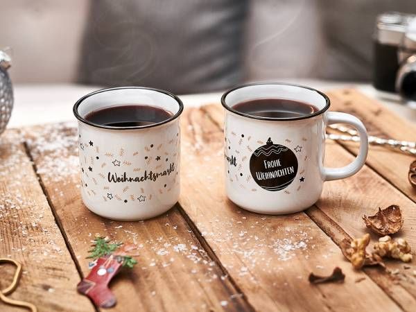 Zwei weiße Emailletassen mit schwarzem Rand, gefüllt mit dunklen Getränken, stehen auf einer hölzernen Oberfläche zusammen mit weihnachtlicher Dekoration und Kunstschnee. Eine Tasse trägt den Schriftzug 'Weihnachtsmarkt', die andere 'Frohe Weihnachten'.