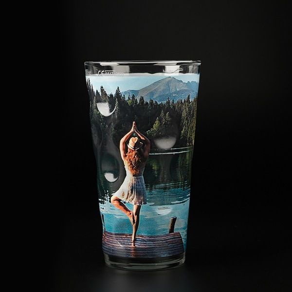 Trinkglas mit aufgedrucktem Bild einer Frau in Yoga-Pose am See vor Bergkulisse.