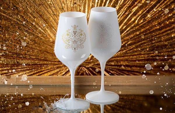 The shot features two stylish wine glasses. The left glass is made of a matte white material and adorned with a delicate, golden pattern reminiscent of stylized grapevines and leaves. The right glass is also white but has a slightly rough, textured surface with tiny, raised dots, giving it a subtle tactile element. Both glasses stand on a glossy, reflective surface that mirrors their shapes and the surrounding light. The background is blurred, showcasing a warm, golden hue with shimmering elements that create a festive or luxurious atmosphere. Several clear water droplets appear to float in the air around the glasses, catching and reflecting the light, which adds a dynamic and fresh touch to the scene.