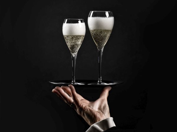 Two filled champagne flutes with a lot of foam on a black tray held by a hand in a dark suit against a black background.