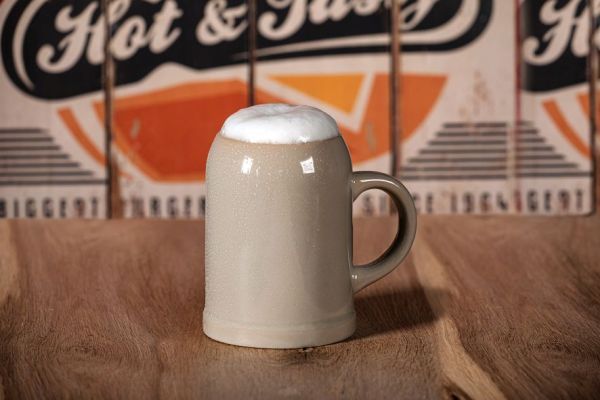 A single, filled stoneware mug with light foam stands on a wooden tabletop in front of a blurred background with lettering.