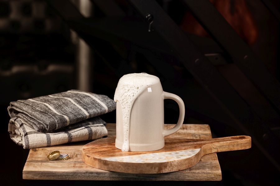 Ein weißer Bierkrug mit überlaufendem Schaum steht auf einem hölzernen Schneidebrett mit zwei Bierdeckeln und gefalteten grauen Geschirrtüchern, vor einem dunklen Hintergrund.