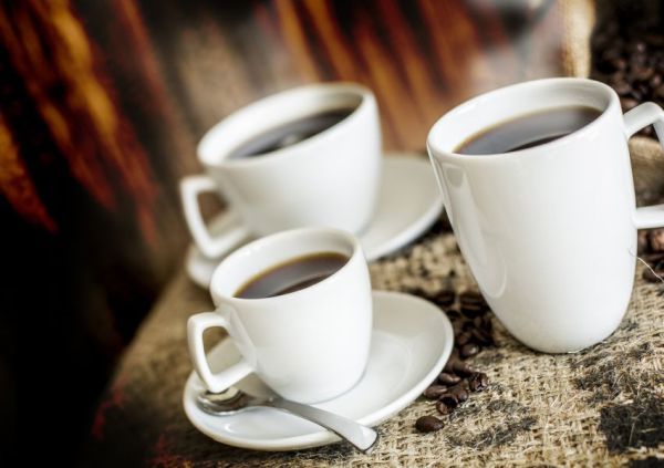 Three white porcelain cups, an espresso cup with saucer and metal spoon, a cappuccino cup with saucer, and a coffee cup are arranged in a triangle on a grayish cloth made of coarse fibers.