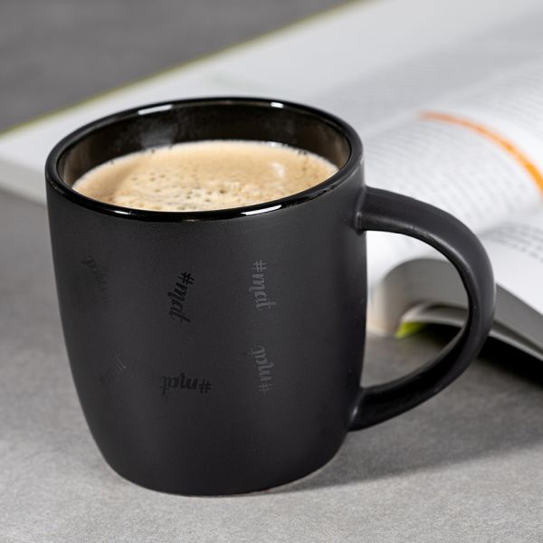 Eine schwarze Kaffeetasse mit hellem Schaum steht auf einer grauen Oberfläche. Mehrere graue Hashtags mit dem Wort 'mat' sind dezent auf der Außenseite der Tasse angebracht. Im Hintergrund ist ein aufgeschlagenes Buch mit unscharfem Text und farbigen Akzenten zu sehen.