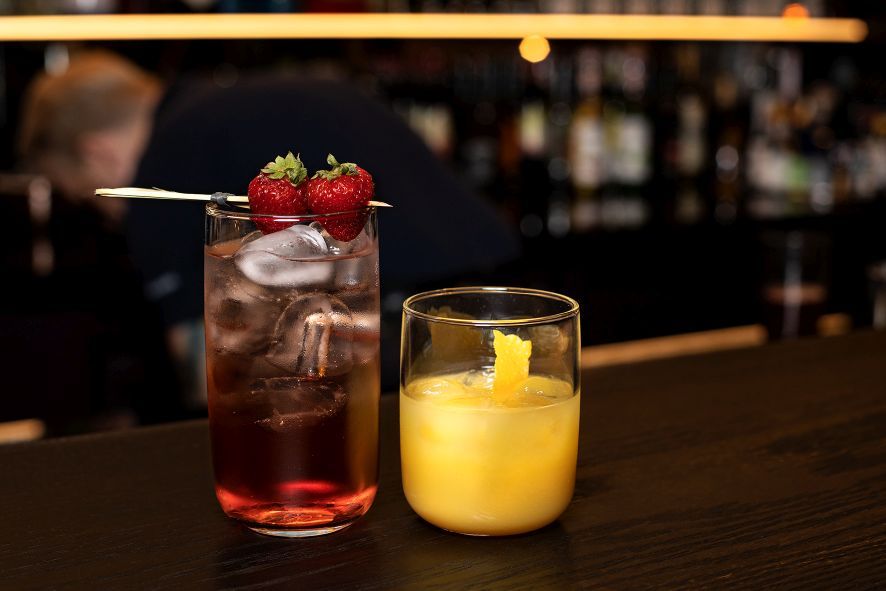 Zwei gefüllte Longdrinkgläser auf einem dunklen Holztisch vor einem verschwommenen Hintergrund einer Bar mit Lichtern und einer Person. Das linke Glas ist rot mit aufgespießten Erdbeeren, das rechte gelb mit einer Orangenzeste.
