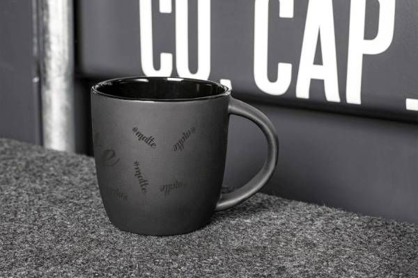 Eine einzelne, matte schwarze Keramiktasse mit Henkel steht auf einer grauen, texturierten Oberfläche vor einer grauen Wand mit weißer Aufschrift '#matte'.
