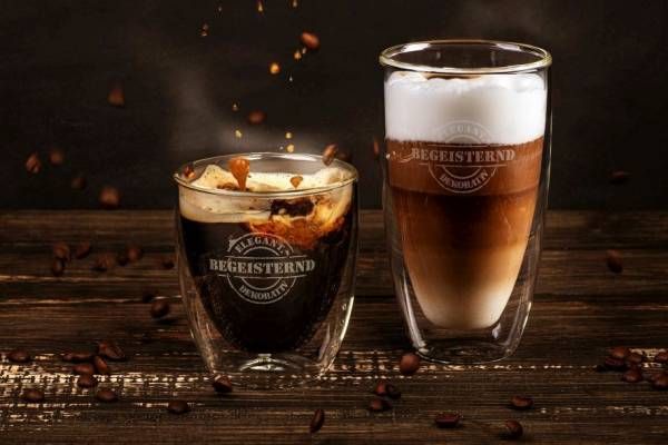 Zwei doppelwandige Glasbecher auf einem dunklen Holztisch vor einem dunklen Hintergrund. Der linke Becher enthält einen Espresso Macchiato, in den gerade Milch gegossen wird, Kaffeebohnen fallen. Der rechte Becher zeigt einen Latte Macchiato mit drei Schichten und Milchschaum.