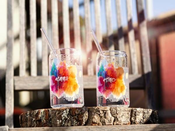 Ein klares Glas in Form einer Getränkedose, aus dem farbige Flüssigkeiten herausspritzen, auf einem weißen Hintergrund.