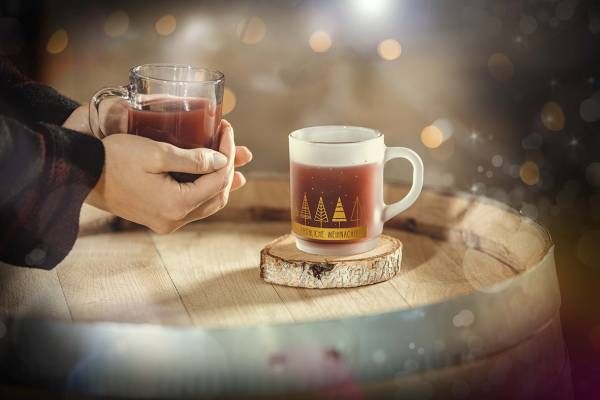 Zwei Hände halten eine klare Glastasse mit einem rotbraunen Getränk über einem Holzfass. Eine weitere, weiße Tasse mit goldenen Tannenbaum-Motiven und dem Schriftzug 'Fröhliche Weihnachten' steht auf einem Holzuntersetzer auf dem Fass. Der Hintergrund ist unscharf mit warmen Lichtern und Schneeflocken-Effekten.