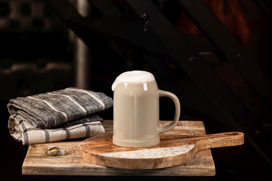 Ein hellbrauner Bierkrug mit überlaufendem Schaum steht auf einem hölzernen Schneidebrett mit zwei Bierdeckeln und gefalteten grau-weiß gestreiften Geschirrtüchern, vor einem dunklen Hintergrund.