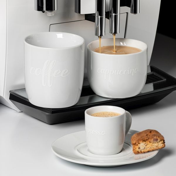 Drei weiße Keramiktassen unterschiedlicher Größen stehen vor einer Kaffeemaschine. Die größte Tasse links trägt die geprägte Aufschrift 'coffee'. Die mittlere Tasse, aus der gerade Kaffee läuft, trägt die Aufschrift 'cappuccino'. Die kleinste Tasse rechts steht auf einer Untertasse und ist mit 'espresso' beschriftet; daneben liegt ein kleines Gebäckstück.