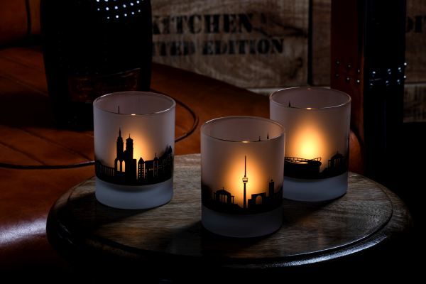 Drei mattierte, zylindrische Teelichtgläser mit schwarzen Skylines verschiedener Städte, beleuchtet durch Teelichter, auf einem dunklen Holztisch.
