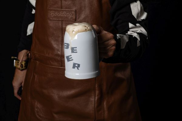 Eine Hand in einem schwarz-weiß gemusterten Pullover hält einen weißen Bierkrug mit der Aufschrift 'BEER' vor einer Person mit einer braunen Lederschürze und dunklem Hintergrund.