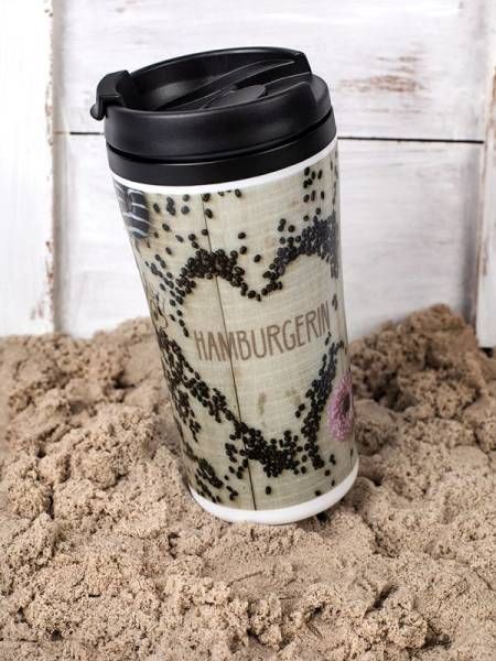 Ein isolierter Reisebecher aus Edelstahl mit einem schwarzen Deckel steht auf sandigem Untergrund vor einer verwitterten Holzwand. Der Becher ist mit einem hellen Muster und dem Wort 'HAMBURGERIN' in einem herzförmigen Design bedruckt.