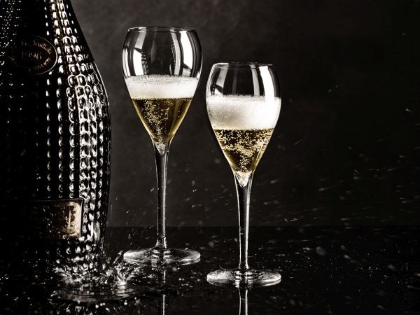 Two filled champagne flutes with foam and a partially visible, dark champagne bottle in the background against a dark background with splashes.