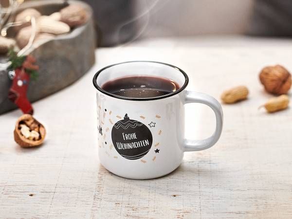 Eine weiße Emailletasse mit schwarzem Rand, gefüllt mit einem dunklen, dampfenden Getränk, steht auf einer hellen Holzoberfläche. Die Tasse ist mit einer Zeichnung einer Weihnachtskugel und dem Schriftzug 'Frohe Weihnachten' verziert. Im Hintergrund weihnachtliche Dekorationen.