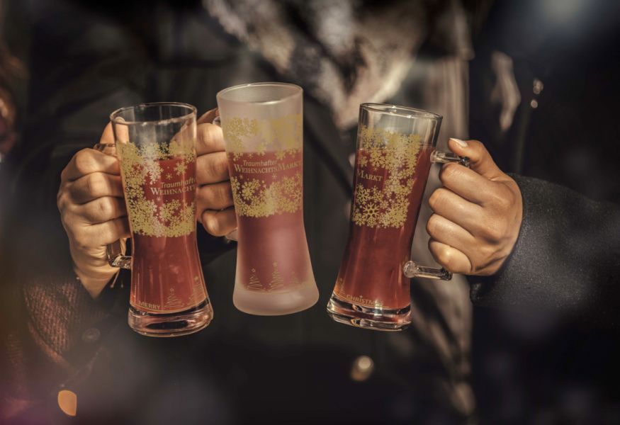 Drei klare Glühweingläser mit Henkel, gefüllt mit rotem Glühwein und dekoriert mit weißen Schneeflocken, gehalten von zwei Personen in dunkler Winterkleidung.