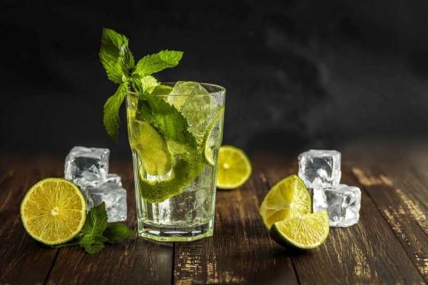 A long drink glass filled with a green drink, ice cubes, lime wedges, and mint on a dark wooden table against a dark background with limes and ice cubes.